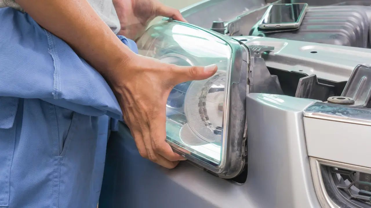 Mechanic in blue uniform installing a clean headlight lens assembly into a silver car's front end